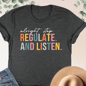May include: A dark heather grey t-shirt with the text "alright stop regulate. and listen." in colorful letters.