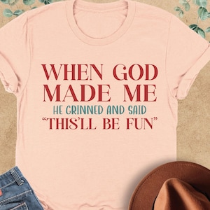 Puede incluir: Una camiseta color melocot&oacute;n con el texto "WHEN GOD MADE ME HE GRINNED AND SAID 'THIS'LL BE FUN'" en rojo. La camiseta se combina con unos vaqueros azules y un sombrero marr&oacute;n. La camiseta est&aacute; doblada sobre una superficie marr&oacute;n claro.