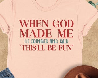 May include: A heather peach t-shirt with the text "WHEN GOD MADE ME HE GRINNED AND SAID 'THIS'LL BE FUN'" in red. The shirt is paired with blue jeans and a brown hat. The shirt is folded on a light brown surface.