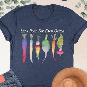 May include: A heather navy blue t-shirt with the text "Let's Root For Each Other" and a graphic of colorful vegetables, including carrots, radishes, and turnips.