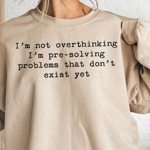May include: A sand-colored crewneck sweatshirt featuring the black printed text: I'm not overthinking I'm pre-solving problems that don't exist yet. The garment has long sleeves with gathered cuffs and a ribbed neckline, presented in a neutral tone.