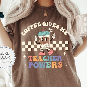 May include: Espresso-colored t-shirt with the text "Coffee Gives Me Teacher Powers" and a cartoon coffee cup graphic. The design includes a checkered pattern and colorful text. The shirt has a crew neck.