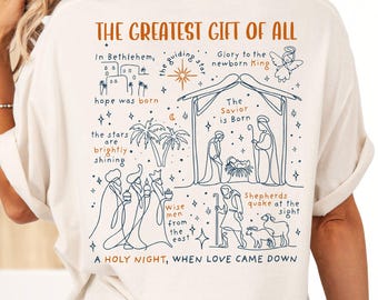 May include: Ivory-colored t-shirt with a blue line drawing on the back. The graphic depicts a nativity scene with text that reads "THE GREATEST GIFT OF ALL" and "A HOLY NIGHT, WHEN LOVE CAME DOWN."