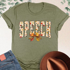 May include: Heather olive green t-shirt with the word "SPEECH" in a fall-themed design. The shirt is paired with blue jeans and a brown hat. The text "HEATHER OLIVE" is in the upper right corner.
