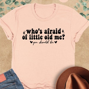 May include: A light pink t-shirt with the text "who's afraid of little old me? you should be" in black. The shirt has a crew neck and short sleeves.
