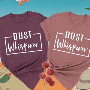 May include: Two heather maroon and heather mauve t-shirts with the text "DUST Whisperer" in a white rectangle on the front. The shirts are laid out on a desert-themed background with a cactus, a hat, and a compass.