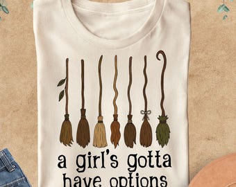 May include: A natural-colored t-shirt featuring a graphic of eight distinct brown and green broomsticks arranged in a row above the black text that reads, a girl's gotta have options. The shirt has a crew neckline and short sleeves.