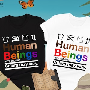 May include: Black and white t-shirts with the text "Human Beings" in various colors. The shirts also have the text "100% Organic Colors may vary." A butterfly and other travel-themed items are in the background.
