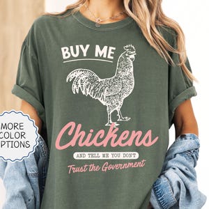 May include: Moss green t-shirt with a white rooster graphic and the text "BUY ME Chickens AND TELL ME YOU DON'T Trust the Government". The shirt has rolled-up sleeves and a relaxed fit. "More Color Options" is also visible.