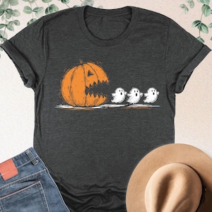May include: Dark grey heather t-shirt featuring an orange pumpkin with a carved mouth and three white ghosts. The shirt is paired with blue jeans and a tan hat. Text on the top right corner reads "DARK GREY HEATHER".