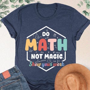 May include: A navy blue t-shirt with the text "DO MATH NOT MAGIC" in a colorful design. The image also shows a pair of blue jeans and a brown hat. The shirt has a crew neck and short sleeves.