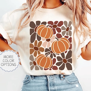 May include: Ivory-colored t-shirt featuring a retro floral and pumpkin design. The graphic includes orange pumpkins and brown and pink flowers. The shirt has the text "MORE COLOR OPTIONS" and "IVORY".