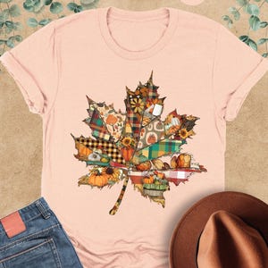 May include: A heather peach short-sleeve t-shirt featuring a large maple leaf graphic composed of various autumn-themed patterns, including plaid, pumpkins, and sunflowers. The shirt is displayed alongside blue denim jeans and a brown fedora hat.