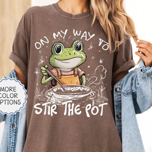 May include: Espresso-colored t-shirt with a whimsical frog graphic stirring a pot, accompanied by the text "ON MY WAY TO STIR THE POT." The design includes a frog wearing an apron, adding a playful touch to the casual wear.