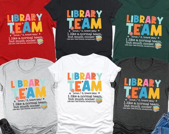 May include: A collection of t-shirts in various colors, including red, black, and green, each featuring the text "LIBRARY TEAM" in colorful letters. The shirts also include a definition and a small graphic of books.