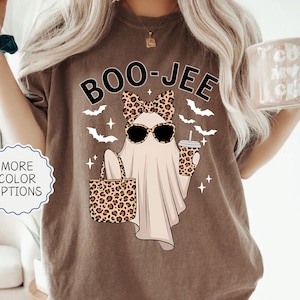May include: Espresso-colored t-shirt with a graphic of a ghost wearing sunglasses and a leopard print bow, holding a leopard print tote bag and a drink. The shirt has the text "BOO-JEE" and is decorated with bats and stars. The shirt is part of the "MORE COLOR OPTIONS" collection.