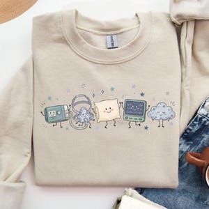 May include: A sand-colored crewneck sweatshirt featuring a graphic print of five anthropomorphic characters: a CPAP machine, a nasal mask, a pillow, a heart rate monitor, and a cloud, all with smiling faces and limbs, surrounded by small stars.
