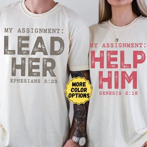 May include: Two off-white short-sleeve t-shirts. The left shirt features brown text reading MY ASSIGNMENT: LEAD HER EPHESIANS 5:23. The right shirt features red text reading MY ASSIGNMENT: HELP HIM GENESIS 2:18. A yellow circular badge says MORE COLOR OPTIONS.