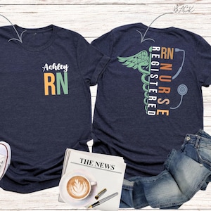 May include: Two navy blue t-shirts with a registered nurse design. The front shirt has the name "Ashley" and the letters "RN" in orange and green. The back shirt has a caduceus with the words "Registered Nurse" and "RN" in orange, green, and blue.