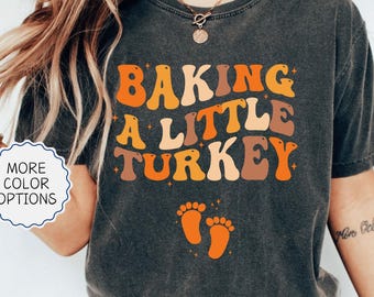 Puede incluir: Una camiseta gris carbón de manga corta con el texto ondulado naranja y marrón BAKING A LITTLE TURKEY y dos pequeñas huellas de pies de bebé naranjas debajo. La camiseta incluye un gráfico circular blanco con MORE COLOR OPTIONS.