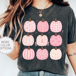 May include: A charcoal gray short-sleeve t-shirt featuring a graphic print of nine pink pumpkins in various patterns, including stripes, polka dots, plaid, leopard print, and breast cancer awareness ribbons, arranged in a three-by-three grid.