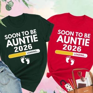 May include: Two t-shirts, one forest green and one red, with the text "SOON TO BE AUNTIE 2026 LOADING..." in white. The green shirt is paired with blue jeans. The red shirt is next to a brown tote bag with accessories.