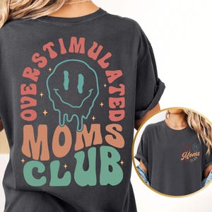 K&ouml;nnte beinhalten: Dunkelgraues T-Shirt mit dem Schriftzug "OVERSTIMULATED MOMS CLUB" in einem Retro-Design. Das Design beinhaltet ein schmelzendes Smiley-Gesicht und Sterne. Das Shirt hat die Farbe Pfeffer.