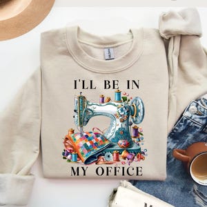 May include: Sand-colored sweatshirt with the text "I'LL BE IN MY OFFICE." The graphic features a detailed illustration of a vintage sewing machine with colorful spools of thread and a quilt. The sweatshirt is paired with blue jeans.