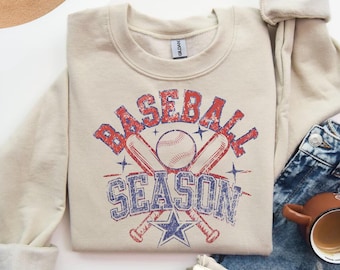 May include: Sand-colored sweatshirt with a distressed "Baseball Season" graphic in red and blue. Features a baseball, bats, and stars. The sweatshirt is folded with denim jeans, a coffee cup, and a newspaper.