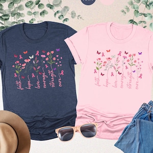 May include: Two t-shirts with a pink floral design and the words "faith, hope, love, courage, strength, cure" printed on them. The shirts are in heather navy and pink.