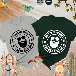 May include: Two t-shirts, one gray and one forest green, each with a circular graphic. The graphic features a bearded face wearing sunglasses and the text "DON'T HATE ME BECAUSE I'M BEARDIFUL". The shirts are short-sleeved.