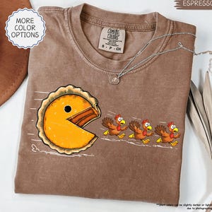 May include: Espresso-colored t-shirt featuring a Pac-Man-style pie chasing three cartoon turkeys. The shirt has a Comfort Colors tag and a silver necklace. The text "MORE COLOR OPTIONS" is in a white circle.