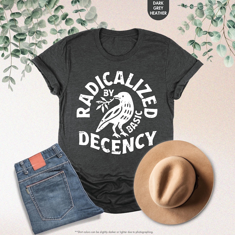 May include: Dark grey heather t-shirt with the text "RADICALIZED BY BASIC DECENCY" in a white circular design with a bird illustration. The image also shows blue jeans and a tan hat.