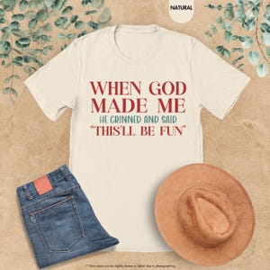 Puede incluir: Camiseta color crema con el texto "WHEN GOD MADE ME HE GRINNED AND SAID 'THIS'LL BE FUN'" en rojo y verde azulado. La camiseta se combina con unos vaqueros azules y un sombrero marr&oacute;n. La camiseta es de material suave.