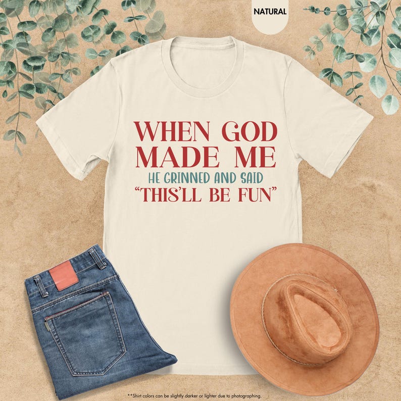 Puede incluir: Camiseta color crema con el texto "WHEN GOD MADE ME HE GRINNED AND SAID 'THIS'LL BE FUN'" en rojo y verde azulado. La camiseta se combina con unos vaqueros azules y un sombrero marr&oacute;n. La camiseta es de material suave.