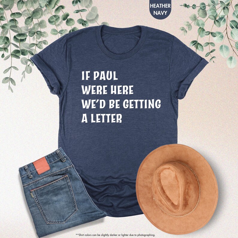 May include: Heather navy t-shirt with the text "IF PAUL WERE HERE WE'D BE GETTING A LETTER". The image also shows a pair of blue jeans and a brown hat. The shirt is made of a soft material.