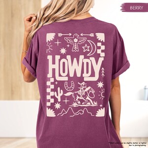 May include: A purple t-shirt with a white graphic design featuring the word "HOWDY" and a cowboy riding a horse. The design also includes various western-themed symbols like stars, arrows, and a horseshoe.