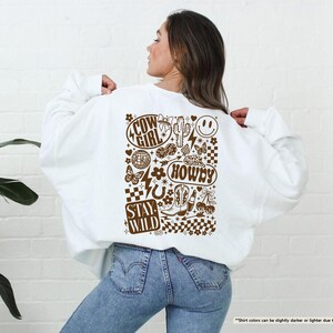K&ouml;nnte beinhalten: Wei&szlig;es Sweatshirt mit braunem Grafikdesign auf dem R&uuml;cken. Das Design enth&auml;lt die W&ouml;rter "COW GIRL", "HOWDY" und "STAY WILD" sowie Bilder von Kaktus, Stiefeln, einem Smiley und anderen Elementen im Western-Stil.