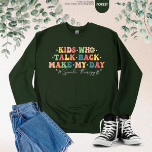 May include: A dark green sweatshirt with the text "KIDS WHO TALK BACK MAKE MY DAY #Speech therapy#" in rainbow colors.