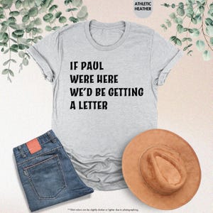 May include: A light gray t-shirt with the text "IF PAUL WERE HERE WE'D BE GETTING A LETTER" in black. The shirt is paired with blue jeans and a brown hat. The words "ATHLETIC HEATHER" are printed at the top.