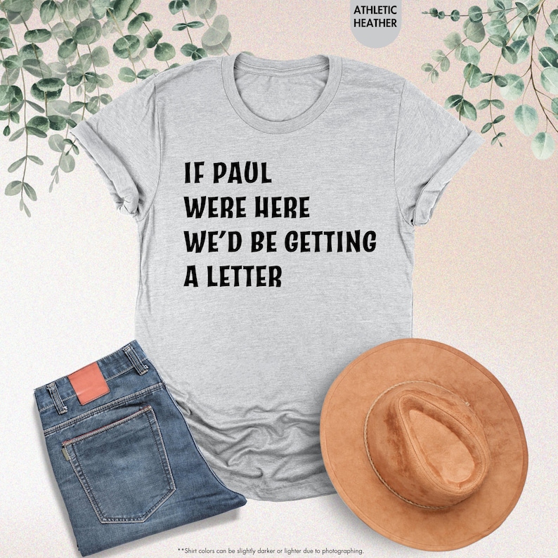 May include: A light gray t-shirt with the text "IF PAUL WERE HERE WE'D BE GETTING A LETTER" in black. The shirt is paired with blue jeans and a brown hat. The words "ATHLETIC HEATHER" are printed at the top.