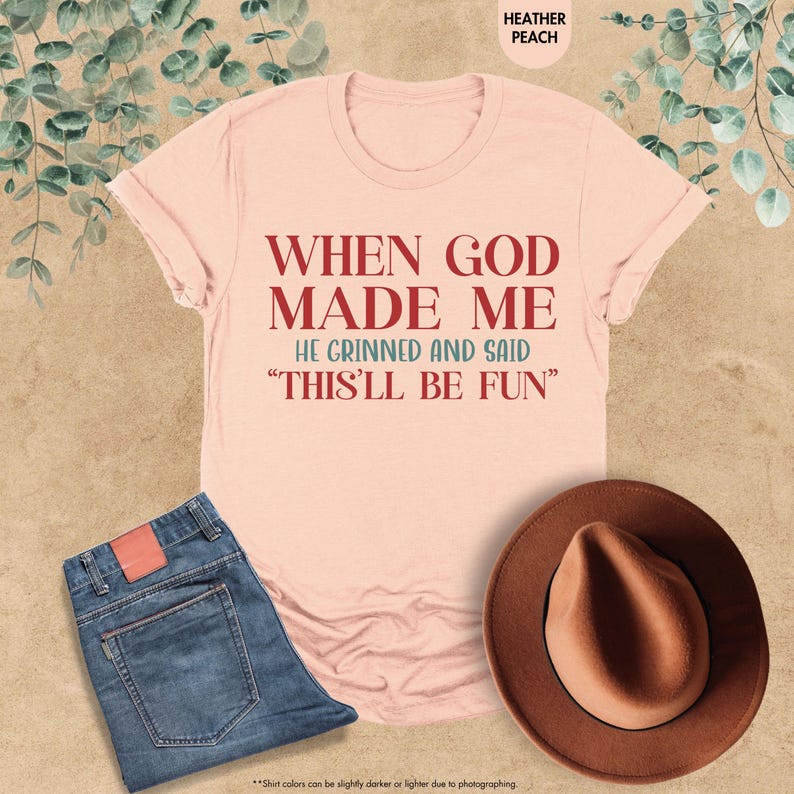Puede incluir: Una camiseta color melocot&oacute;n con el texto "WHEN GOD MADE ME HE GRINNED AND SAID 'THIS'LL BE FUN'" en rojo. La camiseta se combina con unos vaqueros azules y un sombrero marr&oacute;n. La camiseta est&aacute; doblada sobre una superficie marr&oacute;n claro.