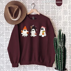 May include: A maroon crewneck sweatshirt featuring a graphic of three white ghosts. The first ghost wears an orange beanie and holds a drink and pumpkin bucket. The second wears a witch hat and holds a broom. The third wears a pink headband.