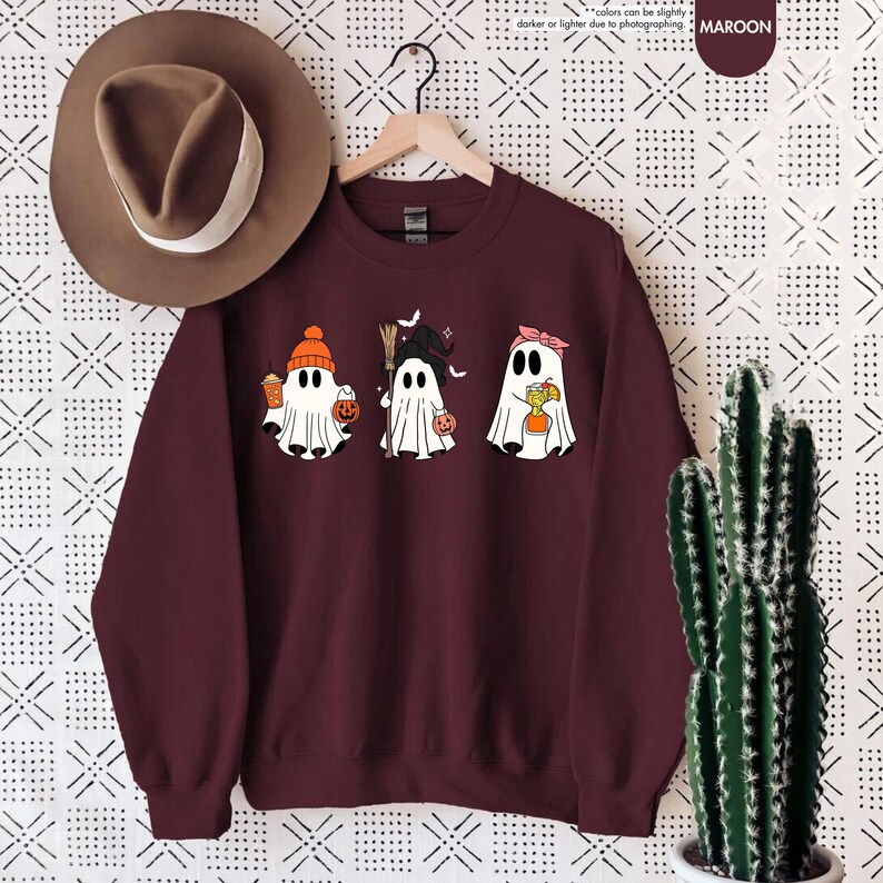 May include: A maroon crewneck sweatshirt featuring a graphic of three white ghosts. The first ghost wears an orange beanie and holds a drink and pumpkin bucket. The second wears a witch hat and holds a broom. The third wears a pink headband.