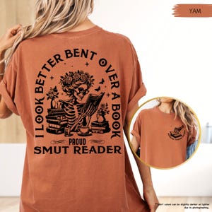 以下が含まれることがあります： さび色のTシャツに、本を読んでいる骸骨の黒いグラフィックが描かれています。Tシャツの文字は「I look better bent over a book, Proud Smut Reader」です。