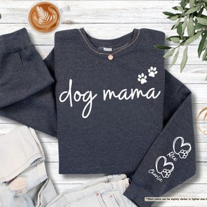 May include: Dark heather gray sweatshirt with the words "dog mama" in white script. The sweatshirt features paw prints and hearts with the names "Rex" and "Charlie" on the sleeve. A cup of coffee sits in the upper left corner.