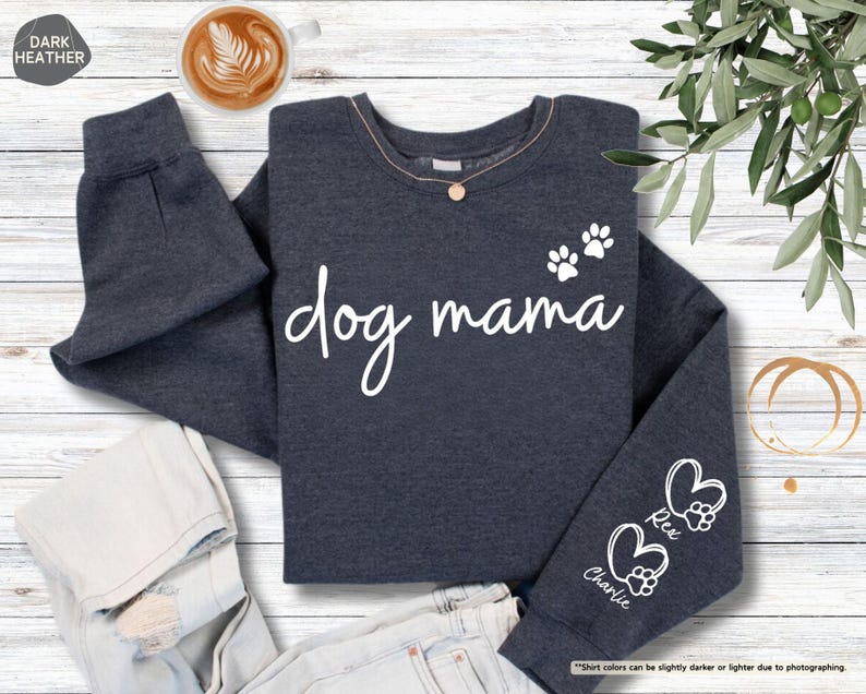 May include: Dark heather gray sweatshirt with the words "dog mama" in white script. The sweatshirt features paw prints and hearts with the names "Rex" and "Charlie" on the sleeve. A cup of coffee sits in the upper left corner.