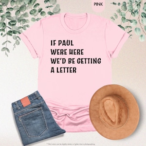 May include: A pink t-shirt with the text "IF PAUL WERE HERE WE'D BE GETTING A LETTER" in black. The shirt is paired with blue jeans and a brown hat. The image also includes greenery in the background.