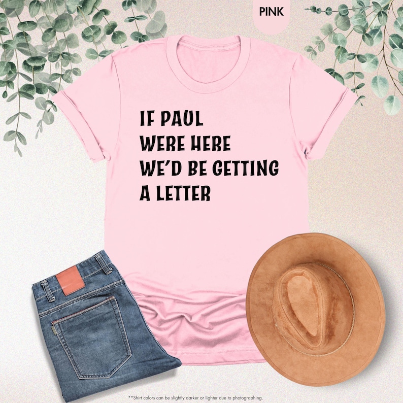 May include: A pink t-shirt with the text "IF PAUL WERE HERE WE'D BE GETTING A LETTER" in black. The shirt is paired with blue jeans and a brown hat. The image also includes greenery in the background.