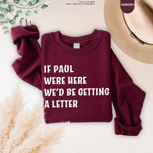 May include: Maroon sweatshirt with the text "IF PAUL WERE HERE WE'D BE GETTING A LETTER" in white. The sleeves are rolled up. The word "MAROON" is in a circle in the upper right corner. The background has a hat and some greenery.